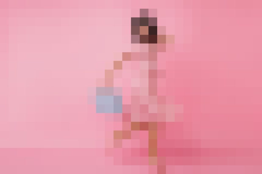 Person wearing a pink outfit stands against a pink background, holding a small blue bag.