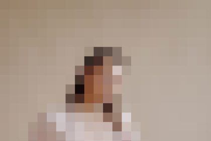 Blurry photo of a person wearing a white dress standing against a beige wall outside a plain room; indistinct details due to pixelation.