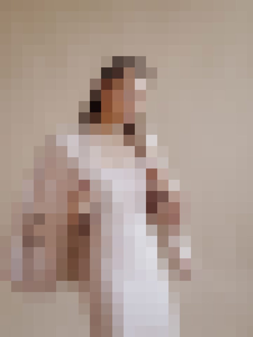 Blurry photo of a person wearing a white dress standing against a beige wall outside a plain room; indistinct details due to pixelation.