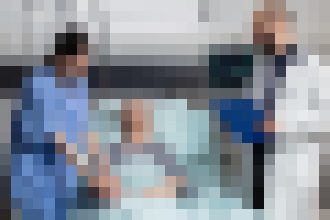 Three healthcare workers stand beside a patient in a hospital bed, suggesting a clinical care scene.