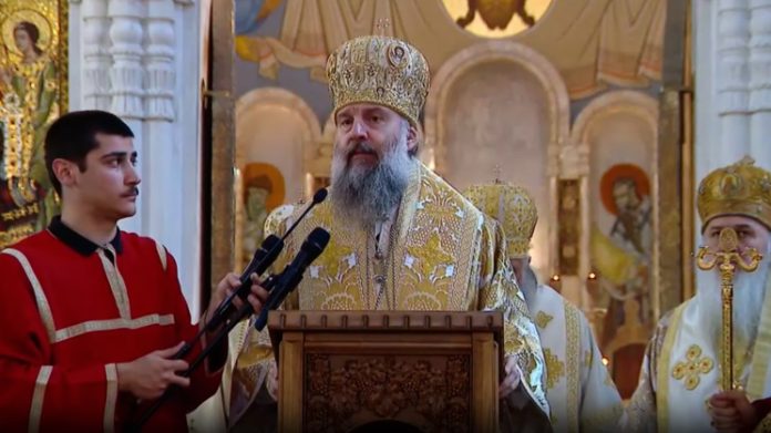 Orthodox bishop in ornate gold vestments delivering a sermon at a wooden pulpit, with a helper holding microphones in a church interior.