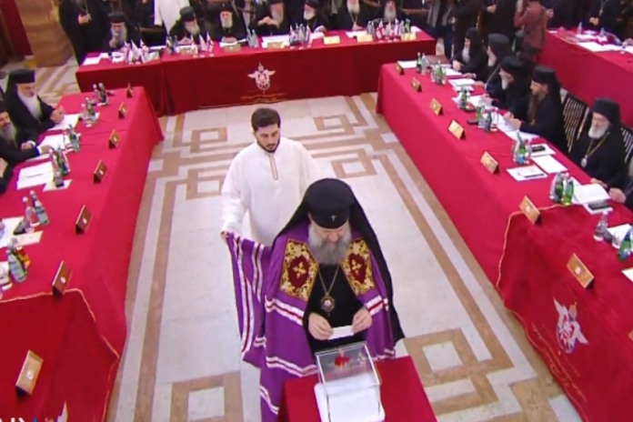 Orthodox priest in ornate purple vestments casts a ballot at a ceremonial voting table surrounded by clergy.