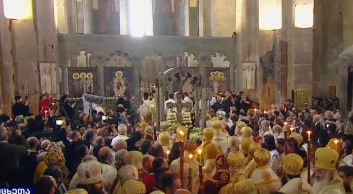 Crowded Orthodox church ceremony with clergy in gold vestments holding candles before the iconostasis.