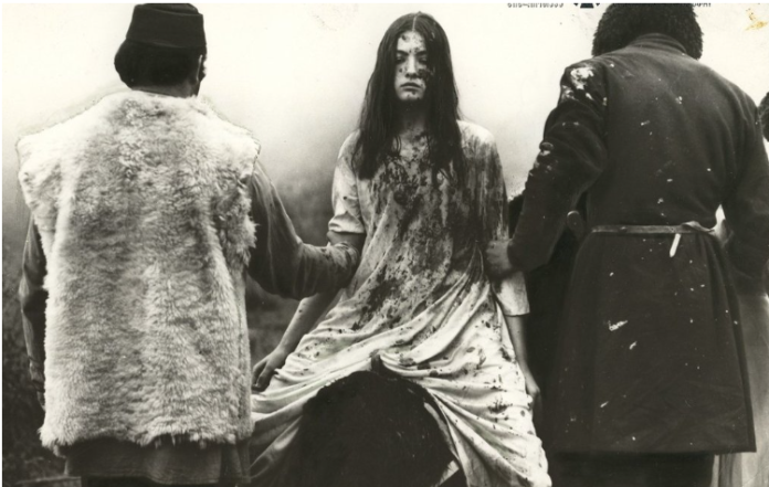 Black-and-white scene: a bloodstained woman in a long gown is being helped by two men in coats, walking away together.