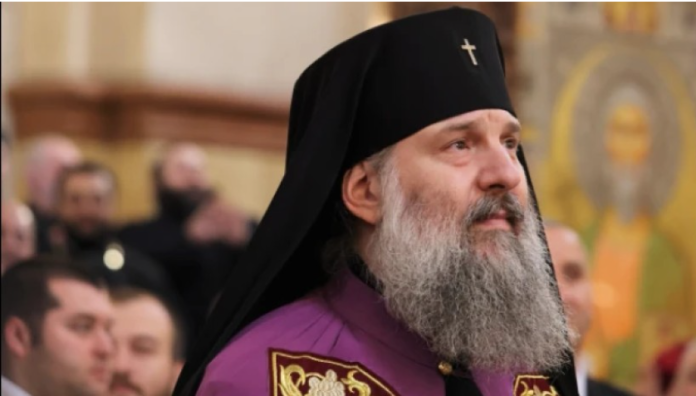 Orthodox priest with a gray beard in purple vestments and a black hat, standing in a church with icons in the background.