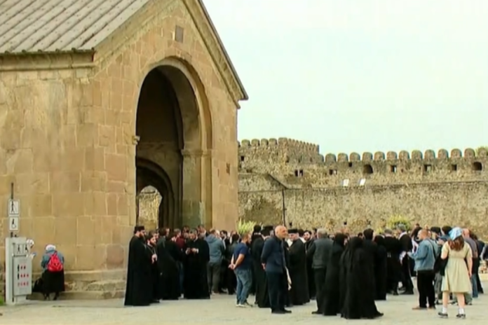 Crowd gathered outside a large stone arch gateway of a fortress, with a crenellated wall in the background.