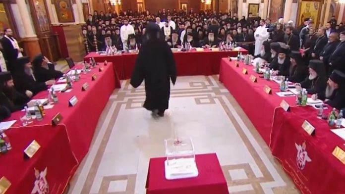 Religious conference with clergy seated around red-draped tables in an ornate hall; a figure in black robes walks toward the front.