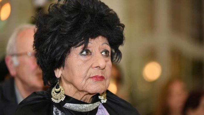 Older woman with a voluminous black hairstyle, chandelier earrings, and red lipstick, looking to the side at a formal event.