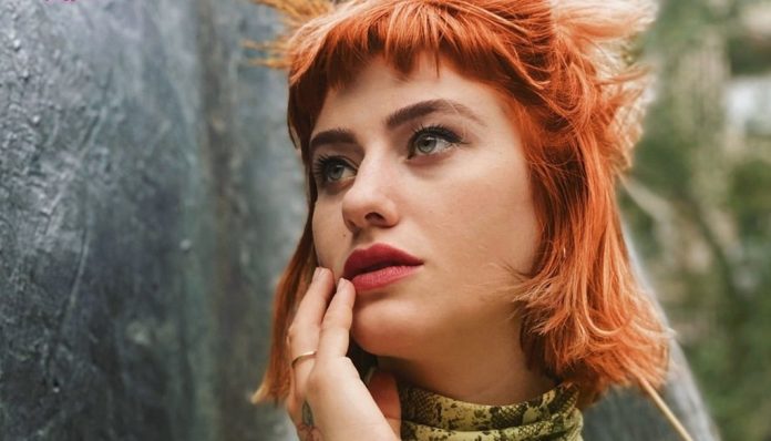 Portrait of a woman with short orange hair gazing upward thoughtfully outdoors near a stone wall