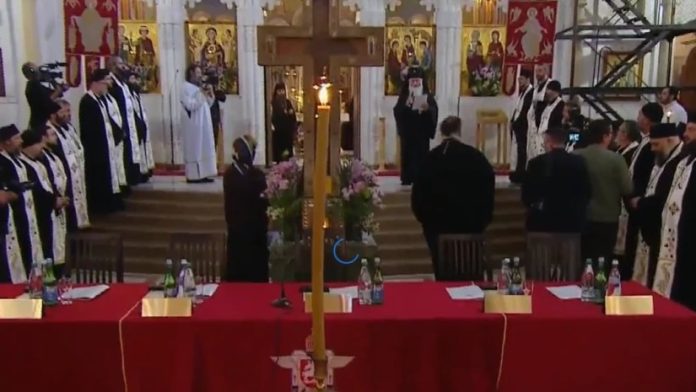 Orthodox ceremony in a church; clergy in black‑and‑white vestments surround a tall central candle with flowers by a red altar table.