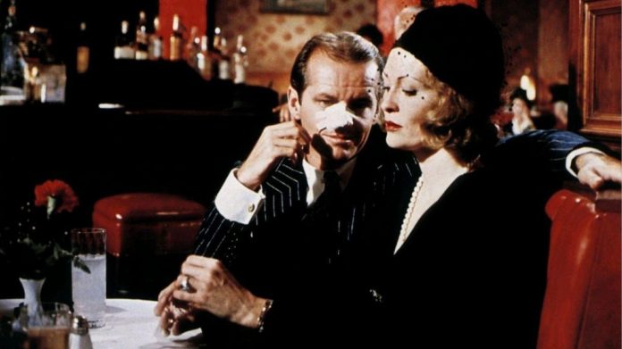 Man with a tissue on his nose sits with a woman wearing a pearl necklace at a retro bar/restaurant.
