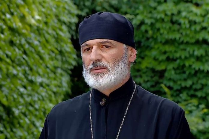 Man with gray beard in a black robe and skullcap, standing outdoors with green foliage behind.