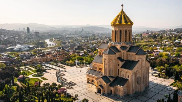 Large brick church with a gold-topped dome and cross, set on a paved plaza in a cityscape with mountains in the distance.