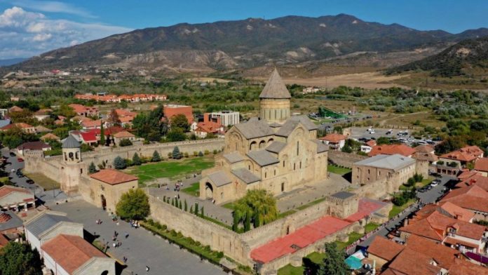 Aerial view of a historic stone church complex with a tall central dome, surrounded by a stone wall and red-roofed houses in a valley. Mountains len mountain range in background.
